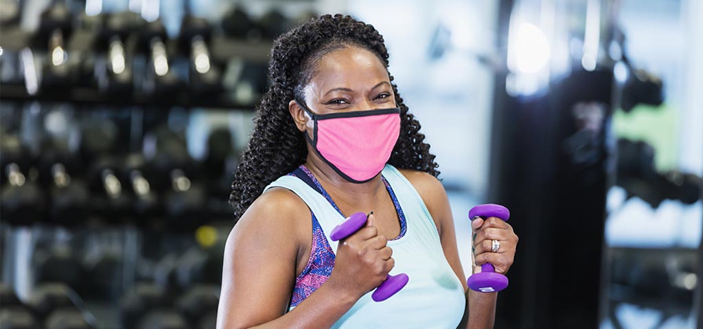 Smiling woman wearing a pink mask, and light blue workout tank top. She is holding 2 purple dumb bells and appears to be in a gym.